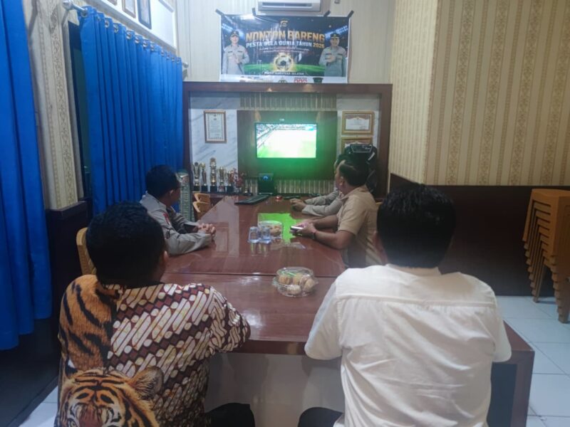 Kapolsek Buay Madang Timur Iptu Swisspo, bersama personel dan warga setempat menyaksikan pertandingan laga Pesta Bola Dunia 2026 antara Amerika Serikat melawan Australia di Mako Polsek Buay Madang Timur, Kabupaten OKU Timur. 