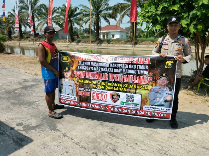 Personel piket Polsek Buay Madang Timur, Aipda Zulkifli dan Bripka Iswanto, membentangkan spanduk maklumat bersama warga saat melaksanakan Giat Sambang dan Sosialisasi Pencegahan Kebakaran Hutan dan Lahan (Karhutla) di wilayah Kecamatan Buay Madang Timur, Kabupaten OKU Timur.